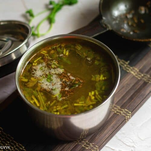 square image of rasam served in stainless vessel placed on a mat with a tempering spoon on the side